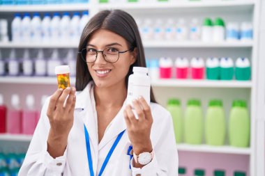 Genç ve güzel İspanyol kadın eczacı gülümseyerek eczanede ilaç şişeleri tutuyor.