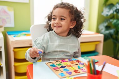 Adorable hispanic girl playing with maths puzzle game sitting on table at kindergarten