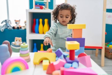 Adorable hispanic girl playing with construction blocks standing at kindergarten