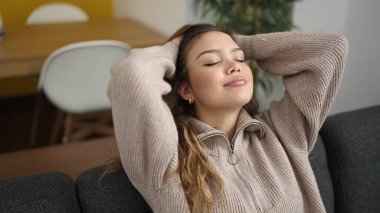 Genç, güzel, İspanyol bir kadın. Elleri başının üstünde, evdeki kanepede oturuyor.