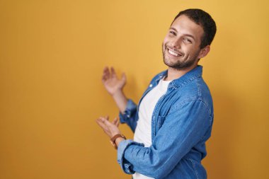 Hispanic man standing over yellow background inviting to enter smiling natural with open hand 