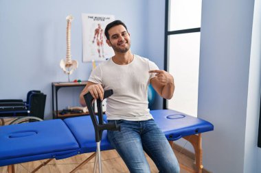 Young hispanic man with beard wearing crutches at rehabilitation clinic pointing finger to one self smiling happy and proud 