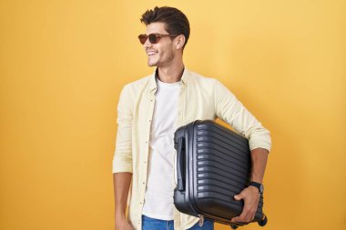 Young hispanic man holding suitcase going on summer vacation looking away to side with smile on face, natural expression. laughing confident. 