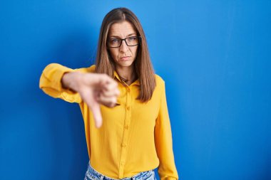 Young woman wearing glasses standing over blue background looking unhappy and angry showing rejection and negative with thumbs down gesture. bad expression. 