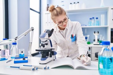 Young blonde woman scientist using microscope writing report at laboratory