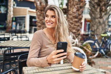 Akıllı telefon kullanan genç sarışın kadın kahve dükkanının terasında kahve içiyor.