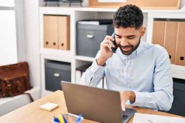 Young hispanic man business worker using laptop talking on the smartphone at office
