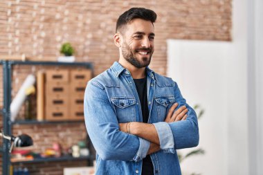 Genç İspanyol adam gülümsüyor. Kollarını kavuşturup sanat stüdyosunda duruyor.