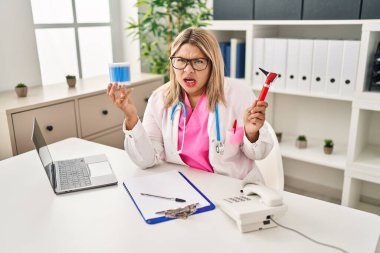 Young hispanic doctor woman holding ear otoscope and cotton buds angry and mad screaming frustrated and furious, shouting with anger. rage and aggressive concept. 