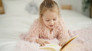 Adorable blonde girl reading book lying on bed at bedroom