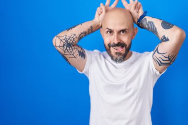 Hispanic man with tattoos standing over blue background posing funny and crazy with fingers on head as bunny ears, smiling cheerful 
