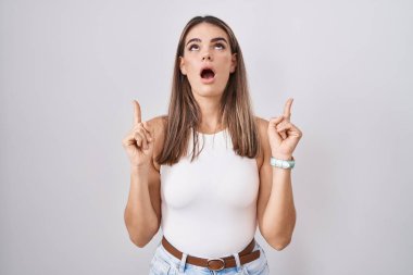 Hispanic young woman standing over white background amazed and surprised looking up and pointing with fingers and raised arms. 
