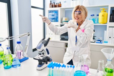Middle age blonde woman working at scientist laboratory pointing aside worried and nervous with both hands, concerned and surprised expression 