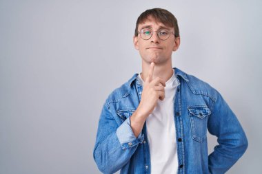 Caucasian blond man standing wearing glasses thinking concentrated about doubt with finger on chin and looking up wondering 