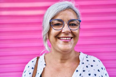 Orta yaşlı bir kadın pembe arka planda kendine güvenerek gülümsüyor.