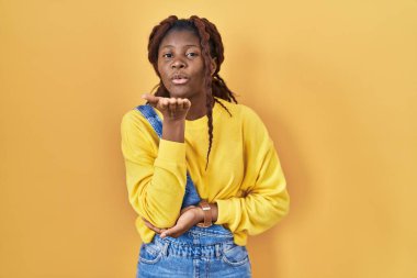 African woman standing over yellow background looking at the camera blowing a kiss with hand on air being lovely and sexy. love expression. 