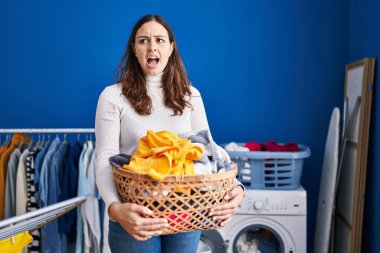 Young hispanic woman holding laundry basket angry and mad screaming frustrated and furious, shouting with anger. rage and aggressive concept. 