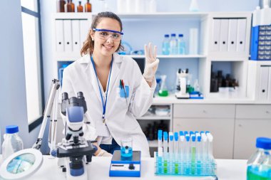 Young hispanic woman working at scientist laboratory showing and pointing up with fingers number three while smiling confident and happy. 