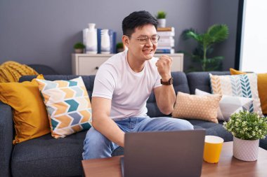 Young asian man using laptop at home sitting on the sofa very happy and excited doing winner gesture with arms raised, smiling and screaming for success. celebration concept. 