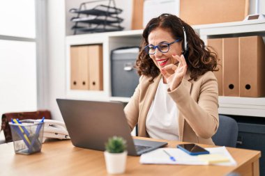 Middle age woman wearing call center agent headset doing ok sign with fingers, smiling friendly gesturing excellent symbol 
