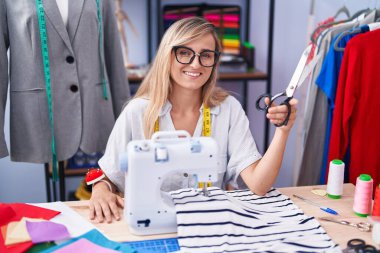 Young blonde woman tailor smiling confident holding scissors at tailor shop