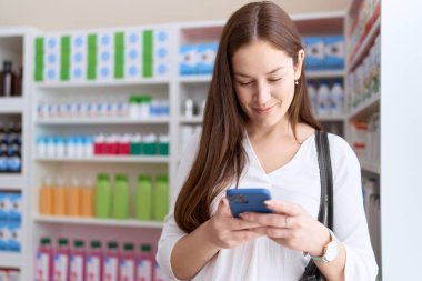 Young beautiful woman customer smiling confident using smartphone at pharmacy