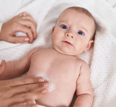 Adorable caucasian baby lying on bed applying cream on body skin at bedroom
