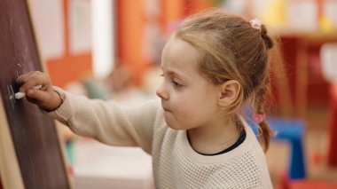 Adorable blonde girl preschool student drawing on blackboard at kindergarten