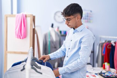 Young hispanic man tailor holding clothes on rack at tailor shop