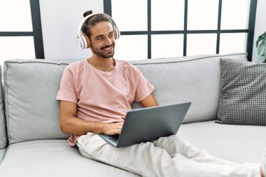 Handsome hispanic man working with laptop sitting on the sofa at the living room at home