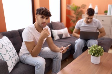 Two hispanic men couple using laptop and smartphone sitting on sofa at home