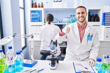 Laboratuvarda çalışan genç adam mutlu ve pozitif gülümsüyor, başparmağıyla mükemmel ve onay işareti yapıyor. 