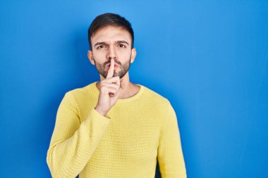 Hispanic man standing over blue background asking to be quiet with finger on lips. silence and secret concept. 