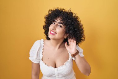 Young brunette woman with curly hair standing over yellow background smiling with hand over ear listening an hearing to rumor or gossip. deafness concept. 