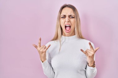 Young blonde woman wearing white sweater over pink background shouting with crazy expression doing rock symbol with hands up. music star. heavy concept. 
