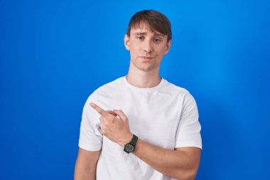 Caucasian blond man standing over blue background pointing aside worried and nervous with forefinger, concerned and surprised expression 