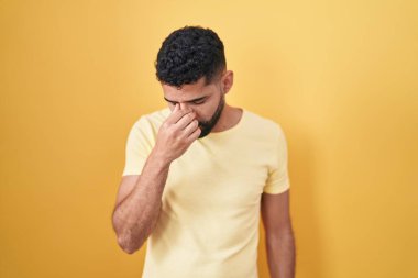 Hispanic man with beard standing over yellow background tired rubbing nose and eyes feeling fatigue and headache. stress and frustration concept. 