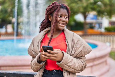 Afro-Amerikalı kadın parkta akıllı telefon kullanarak gülümsüyor.