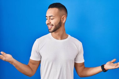 Young hispanic man standing over blue background smiling showing both hands open palms, presenting and advertising comparison and balance 