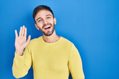 Hispanic man standing over blue background waiving saying hello happy and smiling, friendly welcome gesture 