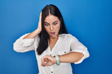 Young hispanic woman standing over blue background looking at the watch time worried, afraid of getting late 