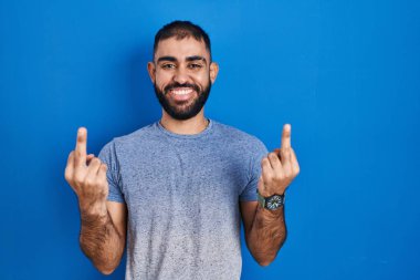 Middle east man with beard standing over blue background showing middle finger doing fuck you bad expression, provocation and rude attitude. screaming excited 