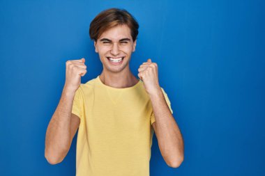 Young man standing over blue background excited for success with arms raised and eyes closed celebrating victory smiling. winner concept. 