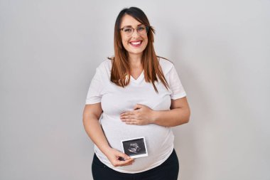Pregnant woman holding baby ecography smiling with a happy and cool smile on face. showing teeth. 