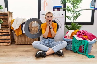 Young blonde woman doing laundry sitting by washing machine afraid and shocked, surprise and amazed expression with hands on face 