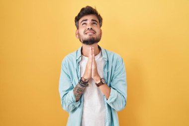 Sarı arka planda dövmeleri olan genç İspanyol bir adam elleri bağlı yalvarıyor ve dua ediyor. Yüzünde umut ifadesi var. Çok duygusal ve endişeli. Yalvarıyor. 