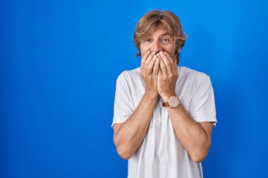 Middle age man standing over blue background laughing and embarrassed giggle covering mouth with hands, gossip and scandal concept 