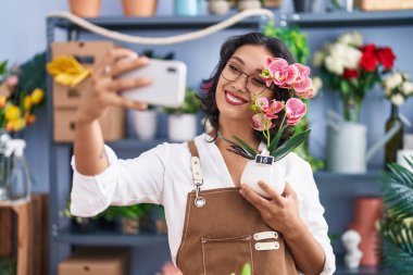 Young beautiful hispanic woman florist make selfie by smartphone holding plant at flower shop