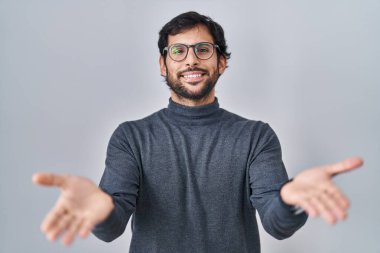 Handsome latin man standing over isolated background smiling cheerful offering hands giving assistance and acceptance. 