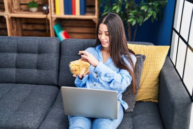 Young beautiful hispanic woman watching movie eating chips potatoes at home